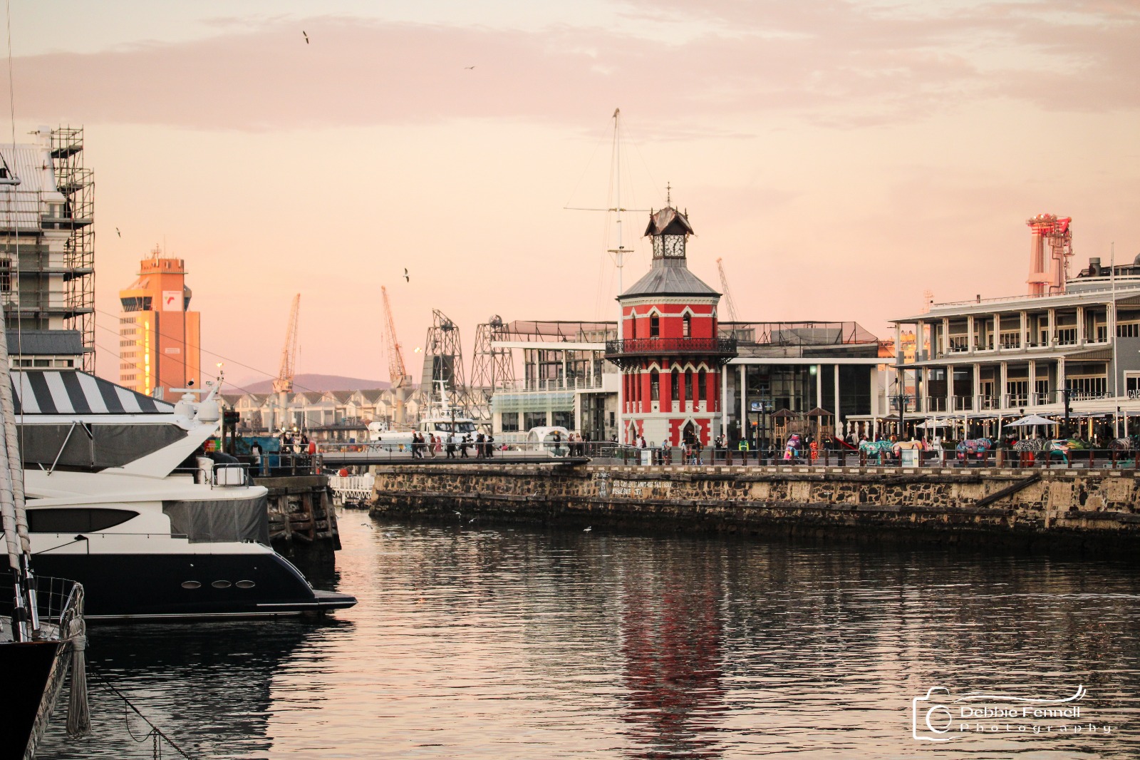 Red Clock Tower viewed across the harbour, with yachts, waterfront buildings and reflections beneath a pink evening sky